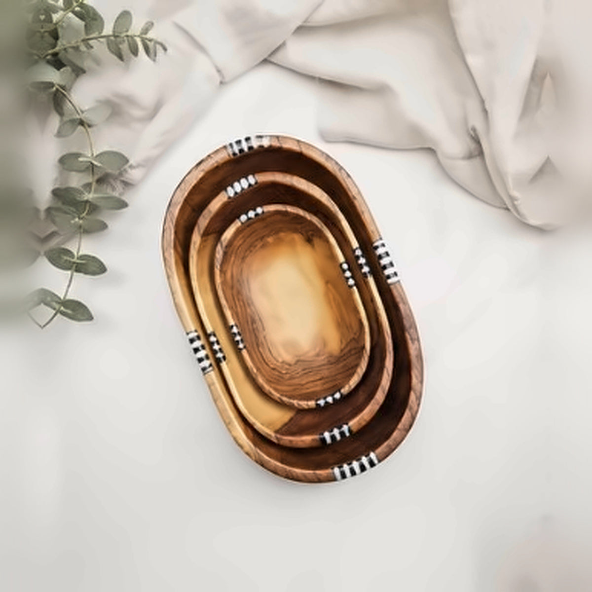 Hand-carved olive wood nesting bowls seen from above on white marble with eucalyptus and linen